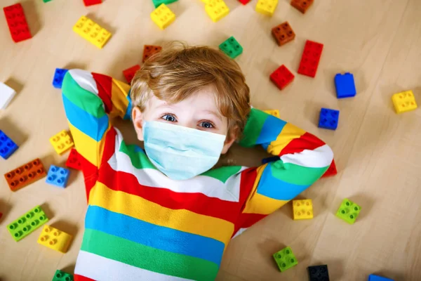 Niño rubio jugando con un montón de bloques de plástico de colores en el interior. Niño pequeño con máscara médica como protección contra la enfermedad de cuarentena por coronavirus pandémico. —  Fotos de Stock