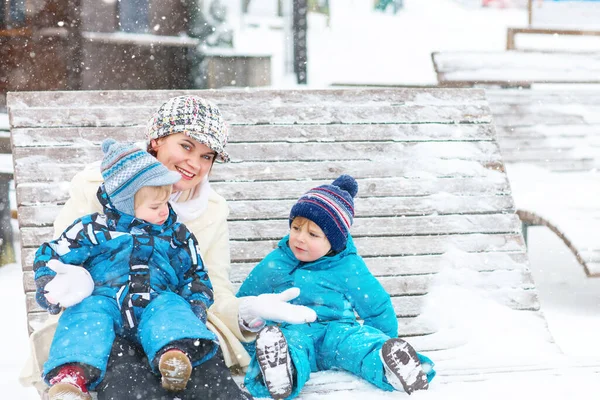 年轻的母亲和两个小孩坐在冬季公园的长椅上。与可爱的小儿子玩耍和拥抱的女人. — 图库照片