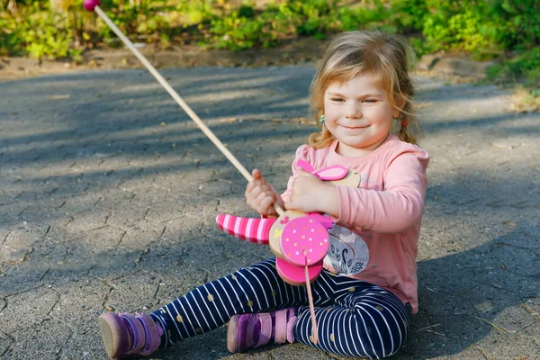 Kleines Mädchen, das an einem sonnigen Frühlingstag im Park spazieren geht. Niedliches entzückendes Kind spielt mit schiebendem Holzspielzeug auf Rädern — Stockfoto