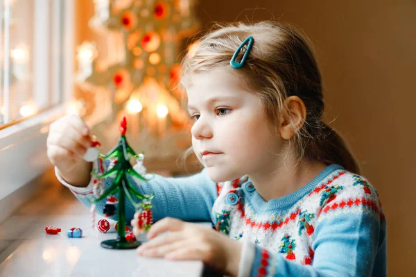 Menina pequena criança sentada à janela e decorando pequena árvore de Natal de vidro com minúsculos brinquedos xmas. A criança sã feliz celebra férias tradicionais de família. Adorável bebê. — Fotografia de Stock