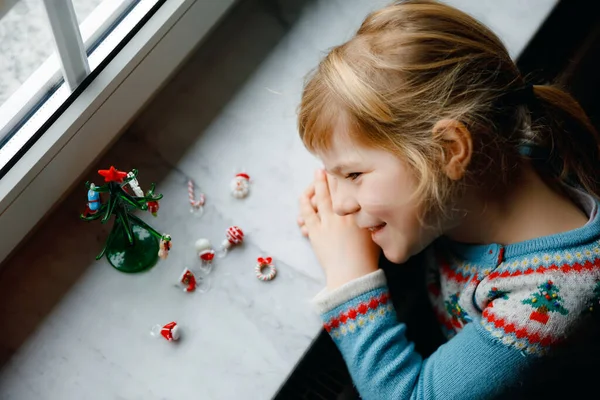 Niña pequeña sentada junto a la ventana y decorando un pequeño árbol de Navidad de vidrio con pequeños juguetes de Navidad. Feliz niño sano celebrar la fiesta tradicional familiar. Adorable bebé.. — Foto de Stock