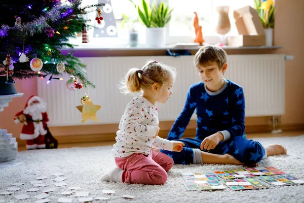 Two little chilren, cute toddler girl and school kid boy playing together card game by decorated Christmas tree. Happy healthy siblings, brother and sister having fun together. Family celebrating xmas — Stock Photo, Image