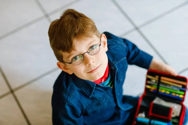 Feliz niño de la escuela buscando un bolígrafo en el estuche. Niño sano con gafas agarrar piensa para las lecciones en la clase elemental. —  Fotos de Stock