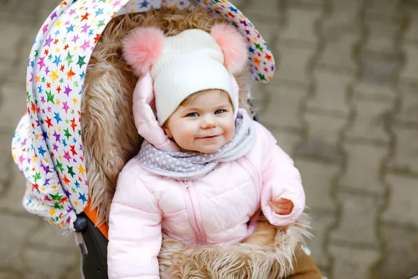 Roztomilá malá krásná holčička sedící v kočárku nebo kočárku na chladném podzimu, zimě nebo na jaře. Šťastné usměvavé dítě v teplých šatech, módní stylový dětský kabát a klobouk. Padající sníh — Stock fotografie