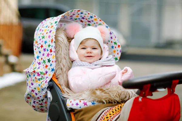 Söt liten vacker liten flicka sitter i barnvagn eller barnvagn på kall höst, vinter eller vår dag. Glad leende barn i varma kläder, mode snygg baby jacka och hatt. Snö faller ner — Stockfoto