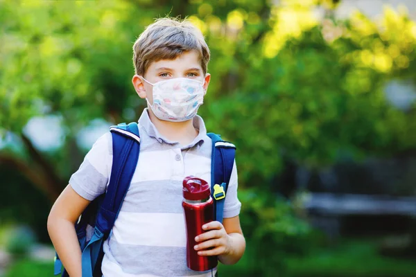 Happy little kid boy, medical mask, water bottle and backpack or satchel. Schoolkid on way to school. Healthy child outdoors. Back to school after quarantine time from corona pandemic disease lockdown — Stock Photo, Image
