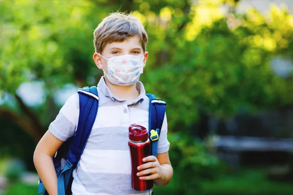 Niño feliz, máscara médica, botella de agua y mochila o mochila. Un colegial de camino a la escuela. Un niño sano al aire libre. Regreso a la escuela después de la cuarentena del bloqueo de la enfermedad pandémica de corona — Foto de Stock