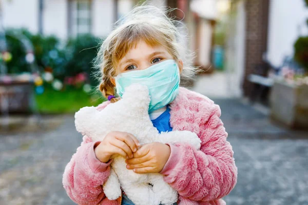 Menina pequena em máscara médica como proteção contra a doença de quarentena do coronavírus pandêmico. Criança bonito usando equipamentos de proteção como luta contra covid 19 e segurando urso brinquedo. — Fotografia de Stock