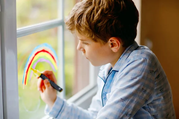 Niño encantador de la escuela en pijama pintando arco iris con colorido color de ventana durante la cuarentena pandémica coronavirus. Niños pintando arcoíris alrededor del mundo con las palabras Vamos a estar todos bien. — Foto de Stock