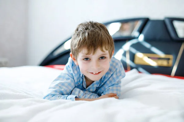 Очаровательный счастливый маленький мальчик после сна в белой кровати в красочной ночной рубашке. Школьник празднует пижамную вечеринку и смотрит в камеру — стоковое фото