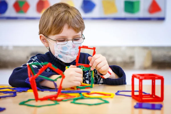 Criança com máscara médica brincando com lotes de blocos de plástico colorido kit no berçário pré-escolar ou escola primária. Equipamento de proteção de uso infantil bonito como luta contra o covid 19 corona vírus. — Fotografia de Stock