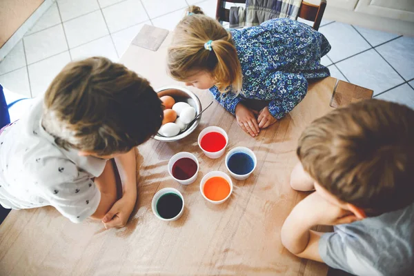 Excited little toddler girl and two kids boys coloring eggs for Easter. Three children, siblings looking surprised at colorful eggs, celebrating holiday with family. From above, unrecognized faces — Stock Photo, Image