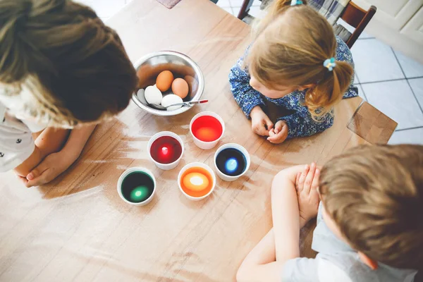 Spännande liten småbarn flicka och två barn pojkar färga ägg till påsk. Tre barn, syskon ser förvånad ut över färgglada ägg, firar semester med familjen. Från ovan, oigenkännliga ansikten — Stockfoto