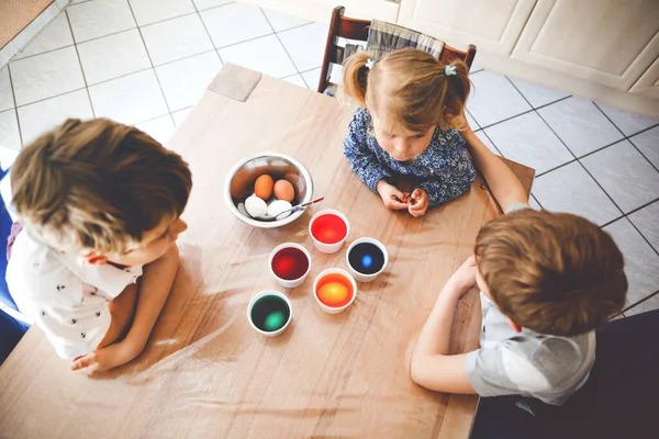 Podekscytowana dziewczynka i dwójka dzieci malujących jajka na Wielkanoc. Trójka dzieci, rodzeństwo wyglądające na zaskoczone kolorowymi jajkami, świętujące wakacje z rodziną. Z góry, nierozpoznane twarze — Zdjęcie stockowe