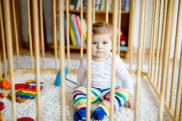 Beautiful little baby girl standing inside playpen. Cute adorable child playing with colorful toys. Home or nursery, safety for kids. Alone baby waiting for mom — Stock Photo, Image