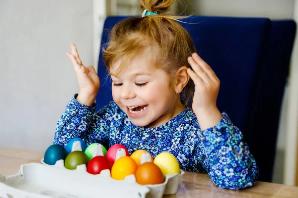 Excited little toddler girl coloring eggs for Easter. Child looking surprised at colored egg hoarding and celebrating catholic and christian holiday with family. Cute kid helping to color, indoors. — Stock Photo, Image