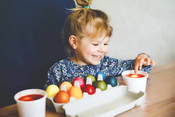 Emocionado menina pequena colorir ovos para a Páscoa. Criança olhando surpreso com ovo colorido acumulando e celebrando férias católicas e cristãs com a família. Criança bonito ajudando a colorir, dentro de casa. — Fotografia de Stock