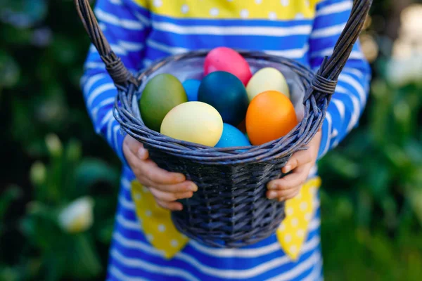 Primer plano de las manos de un niño pequeño sosteniendo la cesta con huevos de colores. Niño divirtiéndose con la tradicional caza de huevos de Pascua, al aire libre. Celebración de la fiesta cristiana — Foto de Stock