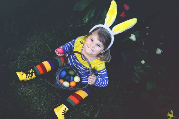 Petite fille mignonne tout-petit avec des oreilles de lapin s'amusant avec des œufs de Pâques traditionnels chassent par une chaude journée ensoleillée, en plein air. Joyeux enfant célébrant des vacances chrétiennes en famille avec panier avec oeuf coloré — Photo