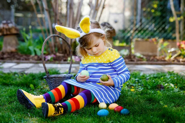 Vacker rolig liten småbarn flicka med kaninöron har kul med traditionella påskägg jakt på varm solig dag, utomhus. Glada barn firar semester. Baby hitta och samla färgade ägg i — Stockfoto