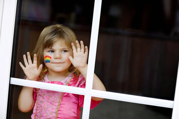 Niedliche einsame kleine Mädchen in Prinzessinnenkleid am Fenster sitzend mit Regenbogen mit bunten Farben im Gesicht während der Coronavirus-Quarantäne. Kinder basteln und bemalen Regenbogen auf der ganzen Welt — Stockfoto