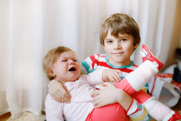 Little kid boy comforts his adorable crying baby sister. Sad toddler girl and lovely brother indoors at home. Girl hit headache. — Stock Photo, Image