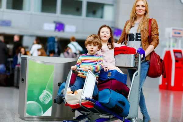 Dvě malé děti, chlapec a dívka, sourozenci a matka na letišti. Děti, rodina cestuje, odlétá na dovolenou letadlem a čeká na vozíku s kufry tlačícími ženy na terminál k letu. — Stock fotografie