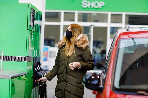 La donna indossa una maschera medica alla stazione di servizio self-service, tiene l'ugello del carburante, ricarica l'auto con benzina durante il blocco pandemico del virus corona. Persone mascherate come misura preventiva e protezione dei merluzzi bianchi — Foto Stock