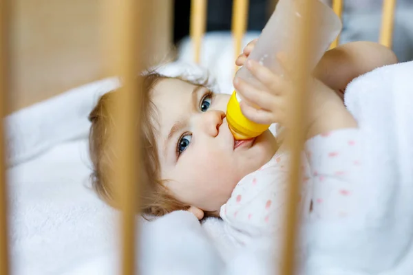 Cute little baby girl holding bottle with formula mild and drinking. Child in baby cot bed before sleeping — Stock Photo, Image