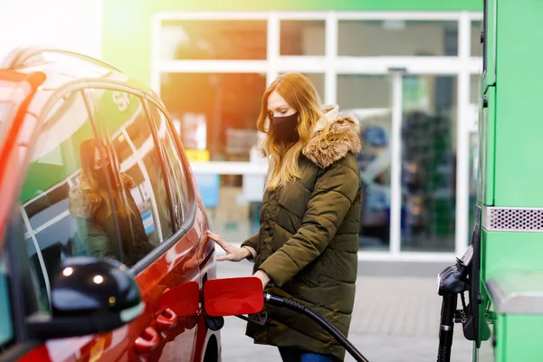 Woman wear medical mask at self-service gas station, hold fuel nozzle, refuel the car with petrol during corona virus pandemic lockdown. People in masks as preventive measure and covid protection