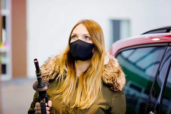 Woman wear medical mask at self-service gas station, hold fuel nozzle, refuel the car with petrol during corona virus pandemic lockdown. People in masks as preventive measure and covid protection