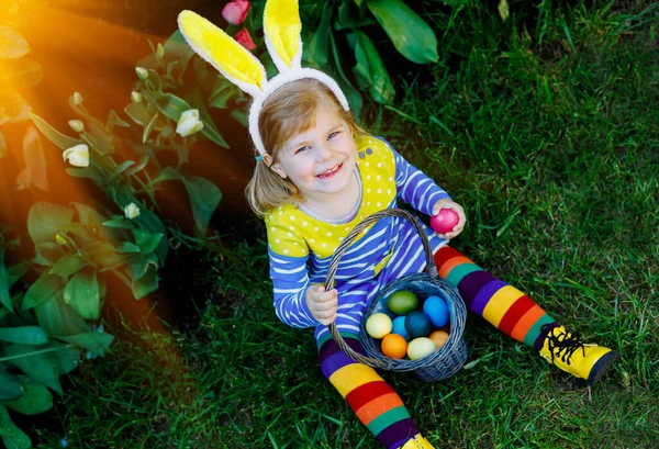 Söt liten småbarn flicka med kaninöron har kul med traditionella påskägg jakt på varm solig dag, utomhus. Glad barn firar familj kristna semester med korg med färgade ägg — Stockfoto