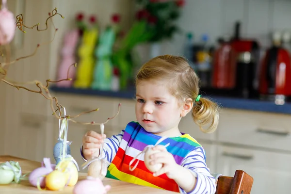 Мила маленька дівчинка-малюк, що прикрашає деревну гілку кольоровими пастельними пластиковими яйцями. Щаслива дитина розважається з великодніми прикрасами. Чарівна здорова усміхнена дитина у сімейному відпочинку — стокове фото