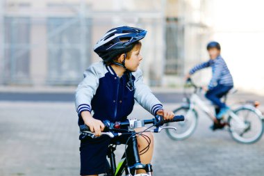 Kasklı iki okul çocuğu şehirde sırt çantalı bisikletle dolaşıyorlar. Renkli giysiler içinde okula bisikletle giden mutlu çocuklar. Çocuklar için güvenli okul yolu.
