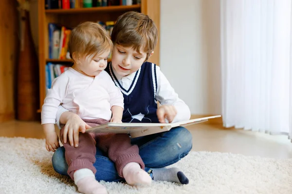 Un garçon de l'école lisant un livre pour une petite fille en bas âge, deux frères et sœurs assis ensemble et lisant des livres. Belle belle famille amoureuse, mignon bébé et enfant s'amuser à la maison, à l'intérieur. — Photo