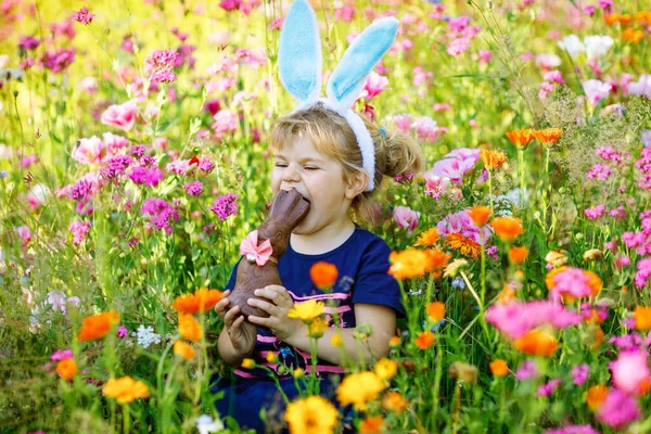 Portriat of adorable, charming toddler girl with Easter bunny ears eating chocolate bunny figure in flowers meadow. Smiling happy baby child on sunny day with colorful flowers, outdoors. — Stock Photo, Image