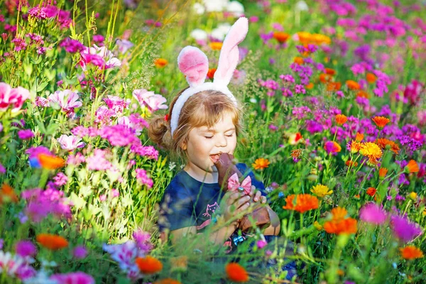 可爱可爱迷人的小女孩，长着复活节兔子的耳朵，在花草甸里吃巧克力兔子的肖像。在阳光灿烂的日子，带着五彩缤纷的花朵，在户外微笑着快乐的孩子. — 图库照片