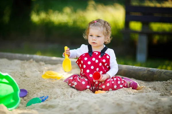 Schattig peuter meisje spelen in zand op buiten speeltuin. Mooie baby in rode gom broek die plezier heeft op zonnige warme zomerdag. Kind met kleurrijk zandspeelgoed. Gezonde actieve baby buiten speelt spelletjes — Stockfoto