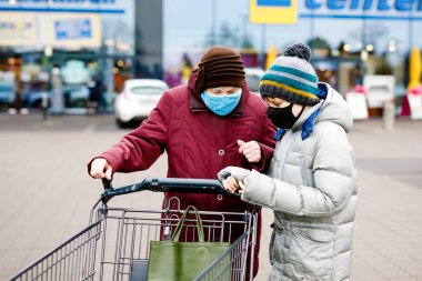 Yaşlı bir kadın ve çocuk salgın koronavirüs hastalığı nedeniyle maske takıyor. Çocuk süpermarkette alışverişte büyükanneye yardım ediyor. Torunum ve büyükannem yük arabasını dışarıda birlikte ittiler.