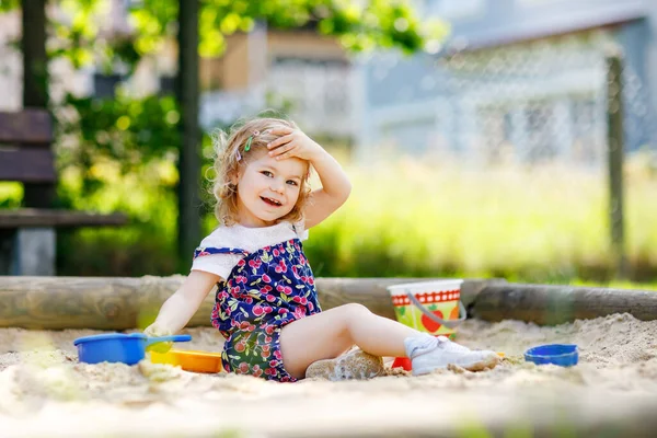 Cute toddler girl playing in sand on outdoor playground. Beautiful baby in red trousers having fun on sunny warm summer day. Child with colorful sand toys. Healthy active baby outdoors plays games — Stock Photo, Image
