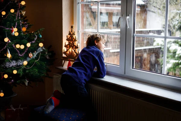 Šťastný rozkošný chlapeček sedí u okna a vánočního stromečku a dívá se ven na sníh na Štědrý den nebo ráno. Usmívající se zdravé dítě fascinované pozorování sněžení a velké sněhové vločky — Stock fotografie