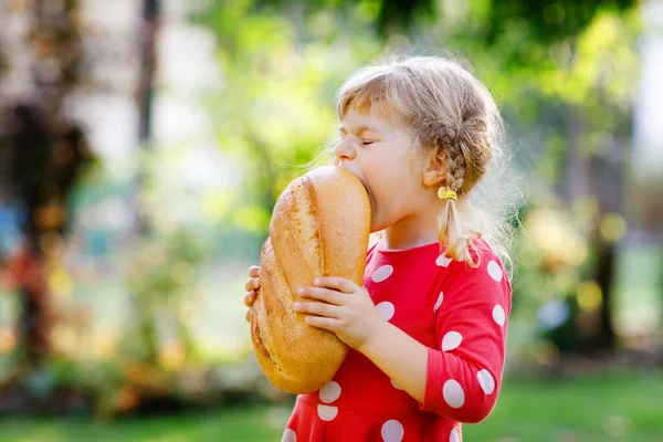 En liten flicka med en stor brödlimpa. Roligt glad barn bita och äta hälsosamt bröd, utomhus. Hungrig unge. — Stockfoto