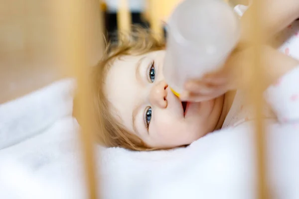 Cute little baby girl holding bottle with formula mild and drinking. Child in baby cot bed before sleeping — Stock Photo, Image
