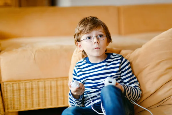 Mignon petit garçon blond avec des lunettes jouant avec une console de jeux vidéo. L'enfant s'amuse à la maison pendant la quarantaine du virus de la couronne. Solitaire garçon seul sans amis, à l'intérieur — Photo