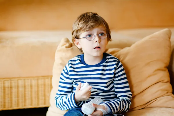 Netter kleiner blonder Junge mit Brille, der mit einer Videospielkonsole spielt. Während der Quarantäne des Coronavirus amüsieren sich Kinder zu Hause. Einsamer Junge ohne Freunde, drinnen — Stockfoto