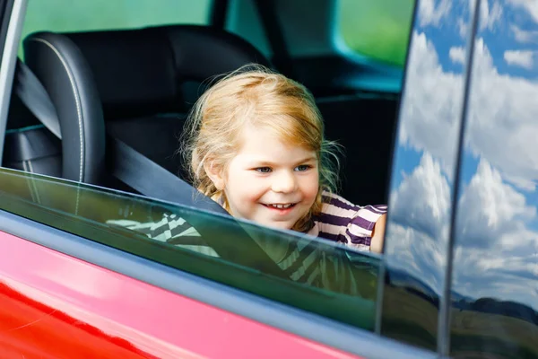 Imádnivaló kisbaba lány ül a gyerekülésen, és néz ki az ablakon a természet és a forgalom. Egy kisgyerek, aki autóval utazik. Gyermekbiztonság az úton. Családi kirándulás és nyaralás nyáron — Stock Fotó