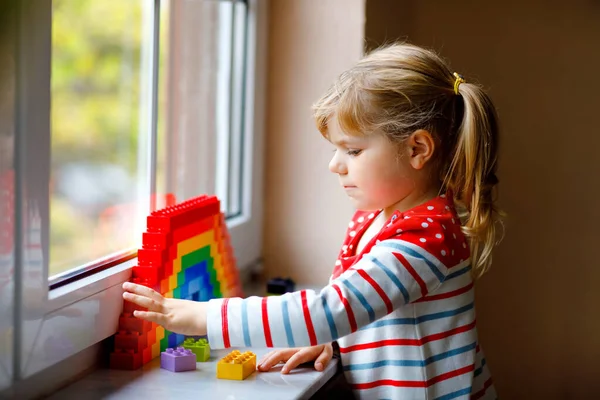 Linda niña pequeña por ventana crear arco iris con bloques de plástico de colores durante la cuarentena pandémica coronavirus. Los niños hicieron y pintaron arcoíris alrededor del mundo como signo. — Foto de Stock