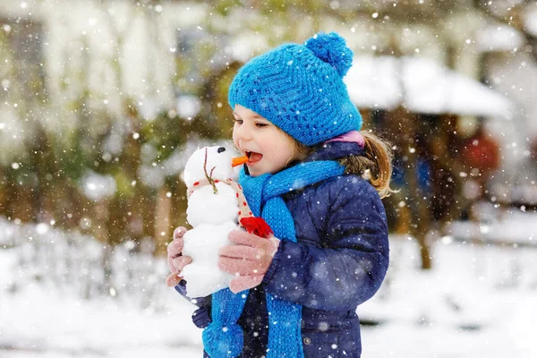 Linda niñita haciendo mini muñeco de nieve y comiendo la nariz de zanahoria. Adorable niño feliz saludable jugando y divirtiéndose con la nieve, al aire libre en el día frío. Ocio activo con niños en invierno —  Fotos de Stock