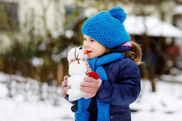 Cute little toddler girl making mini snowman and eating carrot nose. Adorable healthy happy child playing and having fun with snow, outdoors on cold day. Active leisure with children in winter — Stock Photo, Image