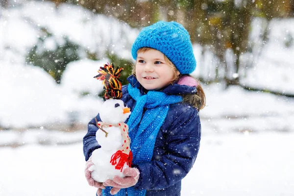 Cute little toddler girl making mini snowman and eating carrot nose. Adorable healthy happy child playing and having fun with snow, outdoors on cold day. Active leisure with children in winter — Stock Photo, Image